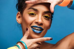 Copa do Mundo: Entre na torcida com a saúde dos olhos!. 2 horizontal stylish mulatto woman with colorful makeup curly hair bun gesturing camera with smile isolated blue wall